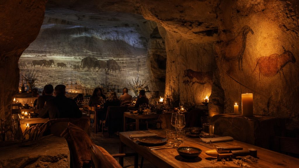 Salle de restaurant immersif à Bordeaux inspirée de la préhistoire, avec parois rocheuses, projections de peintures rupestres et tables élégantes éclairées à la bougie.