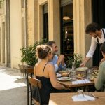 Terrasse d’un restaurant bistronomique chaleureux un dimanche midi, avec un serveur apportant un plat à des clients attablés
