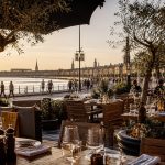 Terrasse de restaurant élégante sur les quais de Bordeaux au coucher du soleil, avec vue sur la Garonne et les façades du centre-ville.