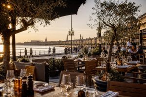 Terrasse de restaurant élégante sur les quais de Bordeaux au coucher du soleil, avec vue sur la Garonne et les façades du centre-ville.